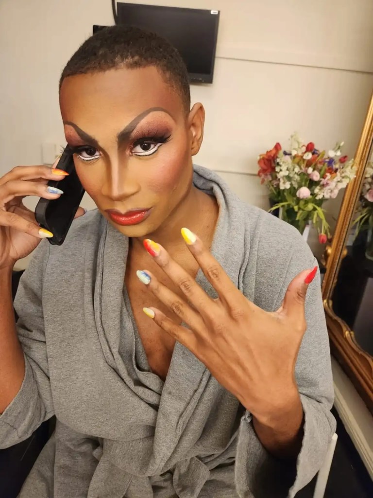 Makeup Artist Spotlight: Kirk Cambridge-Del Pesche: "Don't Give Up! Manifest Your Destiny! Trust Your Process!" 8 A person in dramatic makeup and a gray robe holds a phone to their ear while displaying colorful nail art.