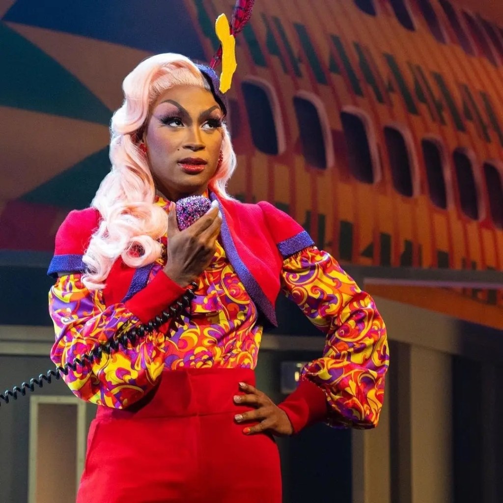 Makeup Artist Spotlight: Kirk Cambridge-Del Pesche: "Don't Give Up! Manifest Your Destiny! Trust Your Process!" 9 A performer in colorful retro attire holds a microphone while standing on stage, with a vibrant backdrop. They have long pink hair and a bold expression.
