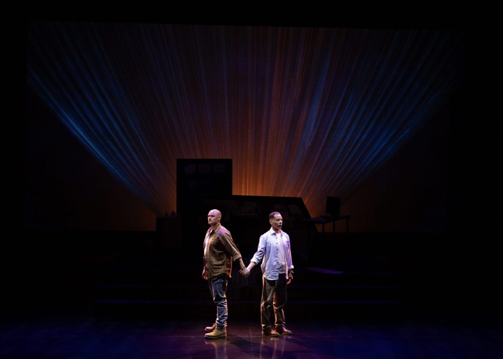 Two male characters standing on a theater stage, holding hands, illuminated by dramatic lighting that creates an emotional atmosphere.