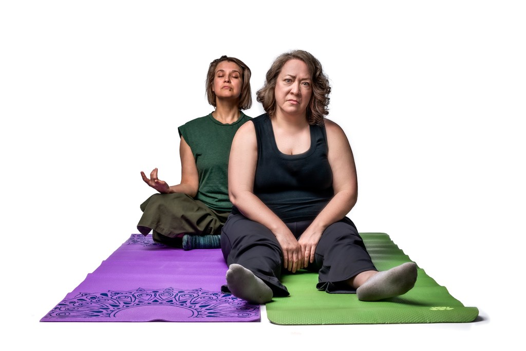 Two women practicing yoga, one seated in a meditative pose on a purple mat, while the other sits on a green mat with a serious expression.