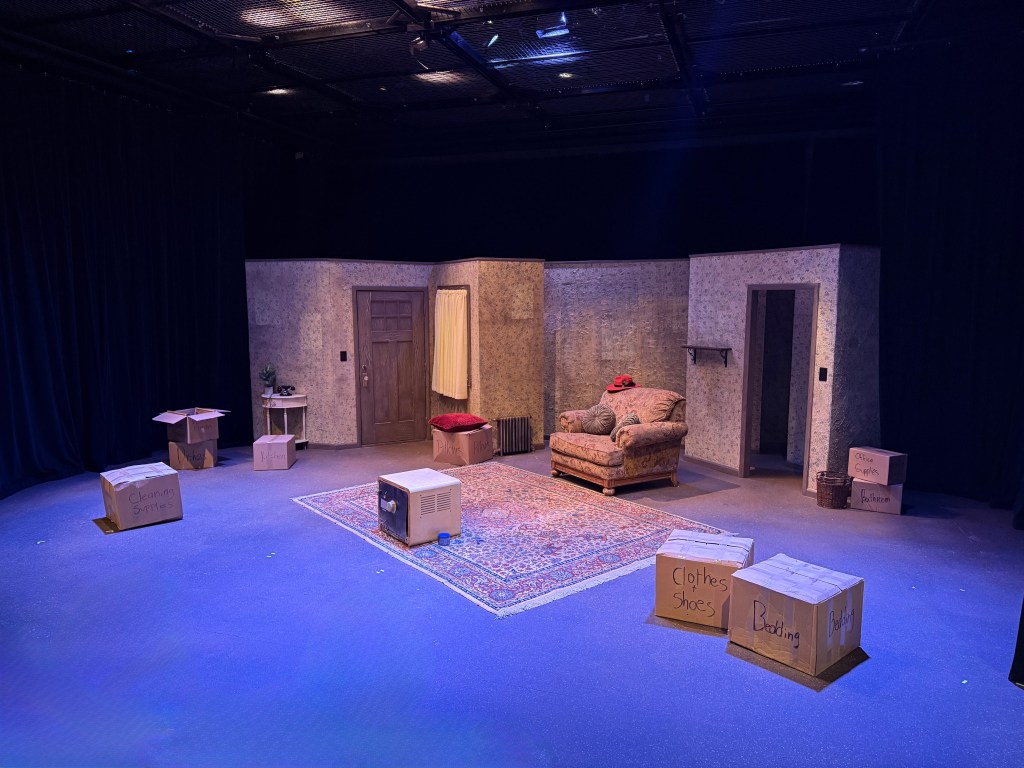 Listening Walls and Lost Names: Westminster University’s "The Hatmaker’s Wife" 2 A stage set for a theatrical production, featuring a modest living room layout with a couch, decorative rug, and various cardboard boxes labeled with items like 'Cleaning Supplies' and 'Clothes'. The walls have a textured finish, and there is a door and window with curtains, creating an intimate and inviting atmosphere.