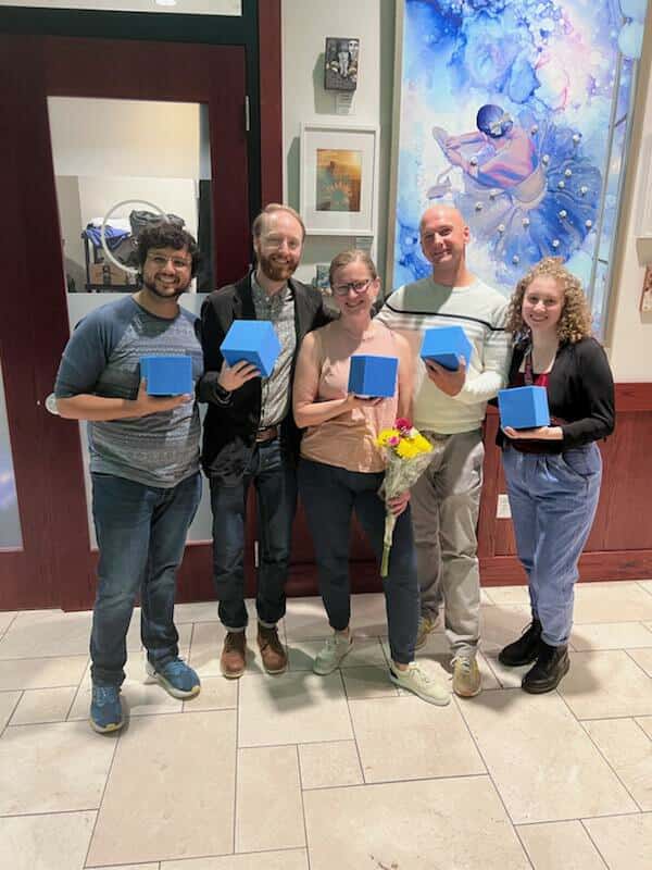 "Book of Ego" by Irene Loy: Drama Therapy and the Excavation of Self 4 A group of five individuals poses together, smiling, while holding blue geometric objects and a bouquet of flowers. The background features a wall with artwork and framed pictures.