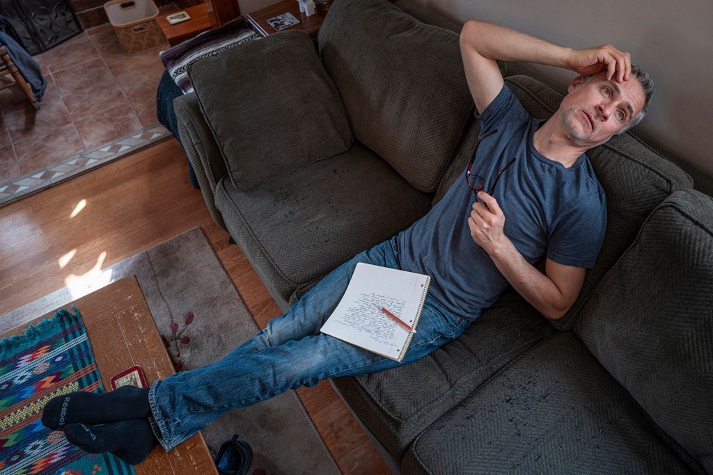 Review: PYGmalion’s “Tiny Beautiful Things” — Writing as Empathy and Witness 2 A man sitting on a couch with a notebook and pen, looking thoughtful and slightly stressed, in a cozy living room setting.