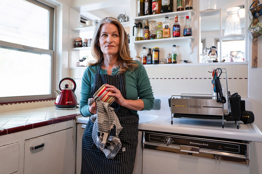 Review: PYGmalion’s “Tiny Beautiful Things” — Writing as Empathy and Witness 7 A woman standing in a kitchen, wearing a black apron over a green sweater, holding a striped cup and a tea towel, with various kitchen items and a kettle in the background.