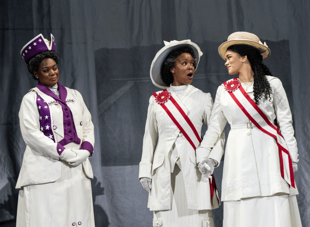 “Keep Marching”: Suffs the Musical Bring the Suffrage Fight into 2025 5 Three women in period costumes representing suffragists, engaged in a moment of emotion on stage, with one woman holding hands with another.