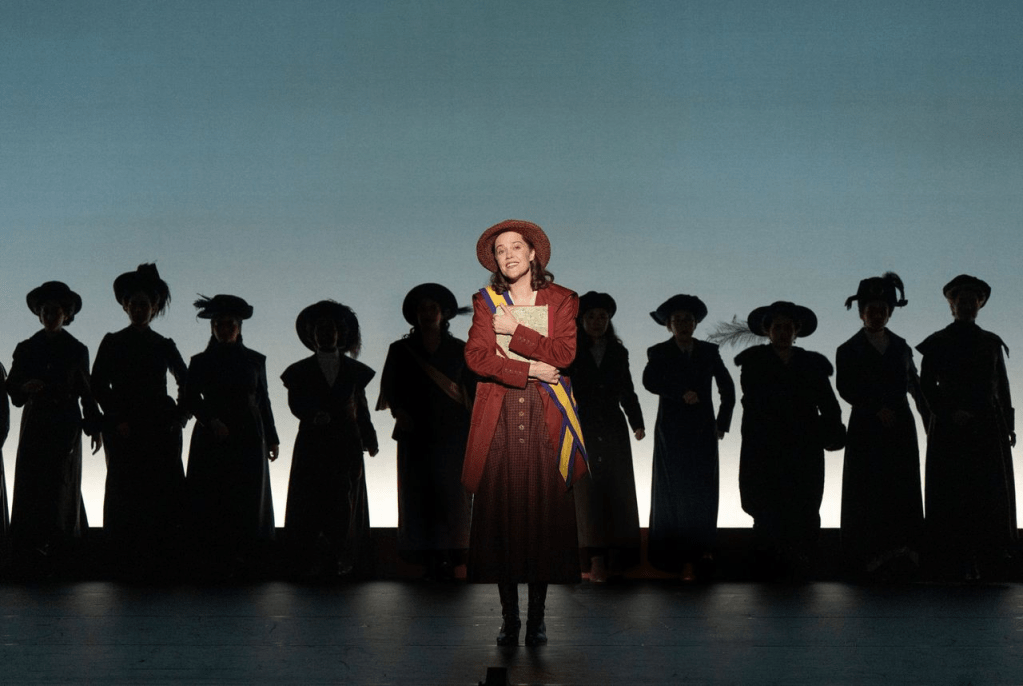 A woman in a red coat and hat stands center stage, holding a book, while silhouettes of other women in historical attire are seen behind her against a soft blue background.