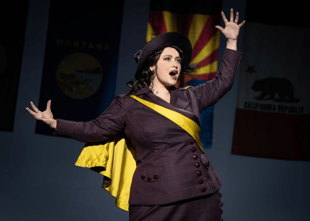 “Keep Marching”: Suffs the Musical Bring the Suffrage Fight into 2025 6 A performer in period costume passionately singing on stage, with flags in the background, portraying a character from the suffrage movement.