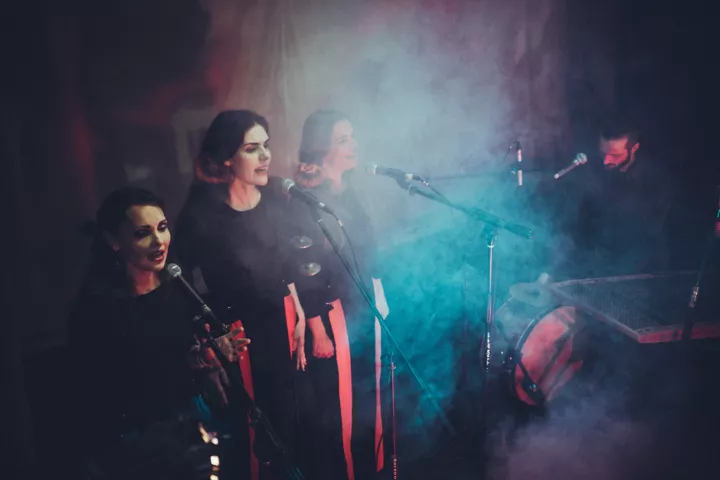 A group of four performers, three women and one man, singing in a smoky environment, with microphones in front of them. The women are dressed in dark outfits with bright accents, while the man plays a traditional instrument.