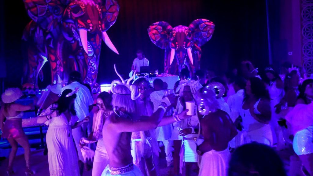SLC White Party 2026: A Voyage Through Time 5 A lively scene at the SLC White Party featuring guests dressed in all white, dancing energetically under purple lighting, with large, colorful elephant decorations in the background.