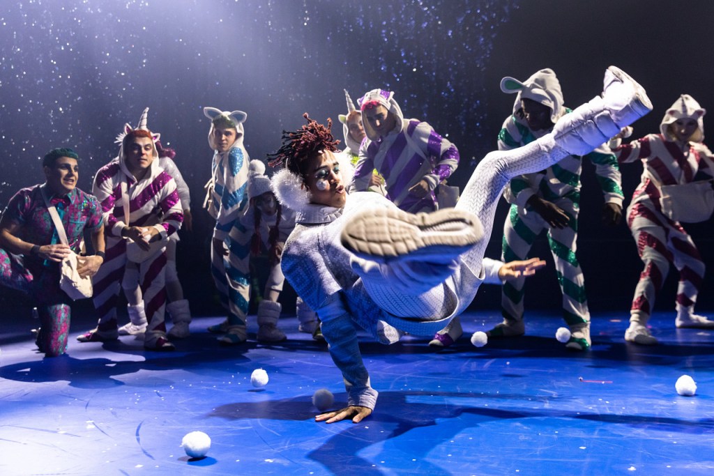 ’Twas the Night Before: Cirque du Soleil’s First Christmas Show Makes Its Salt Lake City Debut 6 A performer dressed in a white costume spins on the ground, surrounded by other dancers in colorful holiday-themed outfits, while snow-like balls are scattered on the stage.