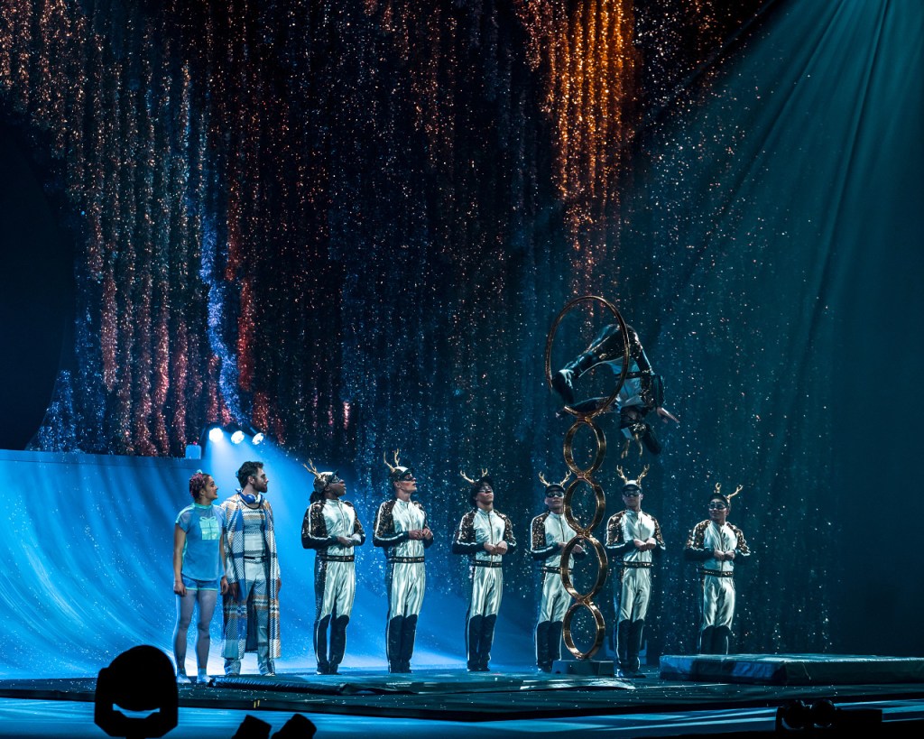 ’Twas the Night Before: Cirque du Soleil’s First Christmas Show Makes Its Salt Lake City Debut 5 A performance from Cirque du Soleil's 'Twas the Night Before,' featuring acrobats in festive costumes and a performer executing a gravity-defying act with rings on stage, surrounded by sparkling lights.