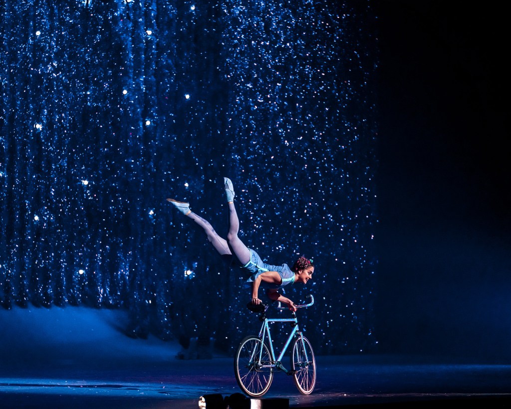 ’Twas the Night Before: Cirque du Soleil’s First Christmas Show Makes Its Salt Lake City Debut 3 A performer showcasing acrobatic skills by balancing on a bicycle, set against a shimmering backdrop of blue lights and falling sparkles.