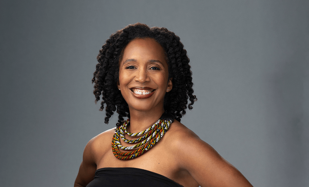Portrait of a smiling woman with natural curly hair wearing a colorful beaded necklace against a gray background.