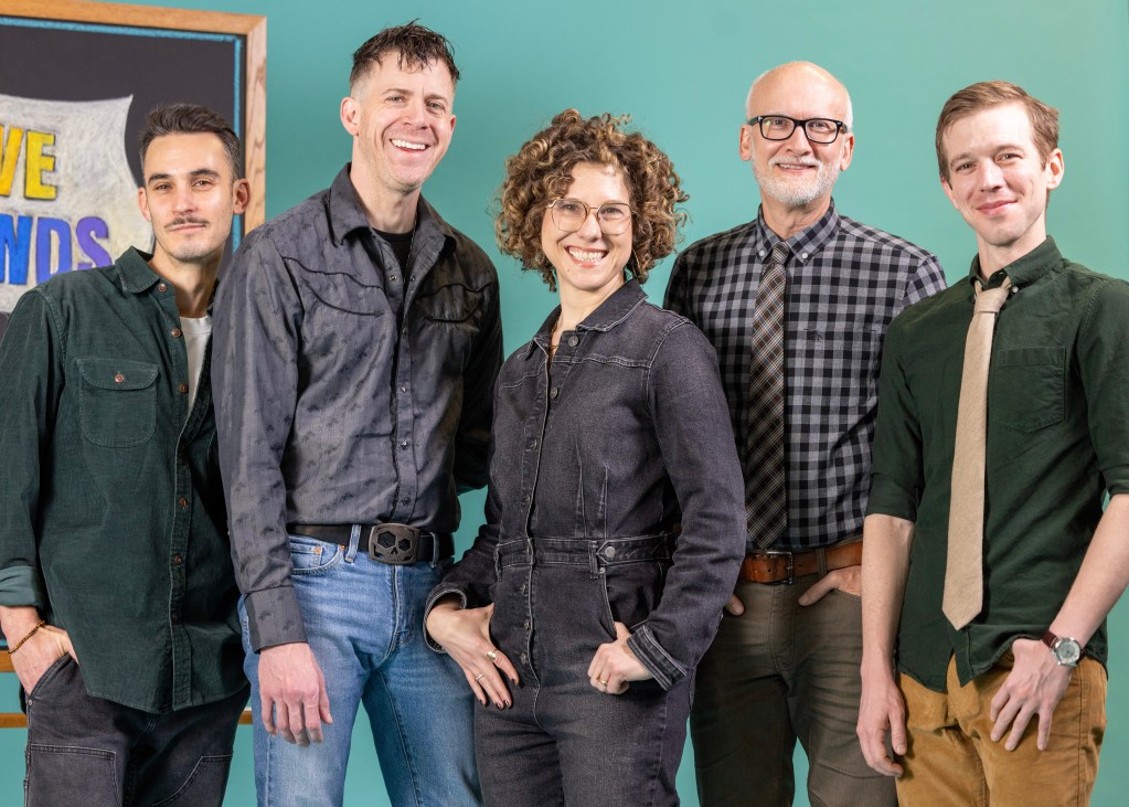 Pioneer Theatre Review: "Ten Brave Seconds" and the Courage of Staying in the Room 6 Five individuals standing together, smiling against a teal background. They are dressed in a mix of casual and semi-formal attire, with two wearing jackets and three in shirts with ties. A chalkboard with text is visible in the background.