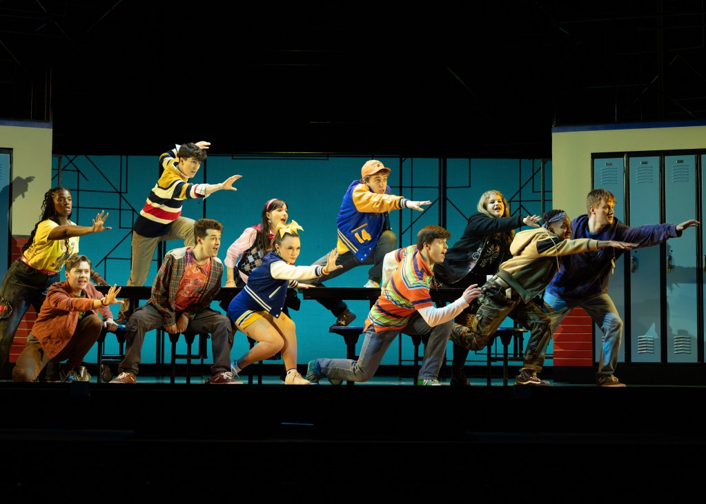 Pioneer Theatre Review: "Ten Brave Seconds" and the Courage of Staying in the Room 4 A group of ten performers striking dynamic poses on stage, wearing colorful costumes, depicting a lively theatrical scene in a school setting.