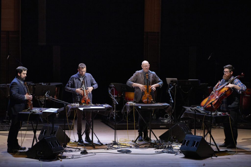 Four musicians performing on stage, each playing string instruments: a violin, a viola, and two cellos, with electronic equipment and microphones around them in a dimly lit concert hall.