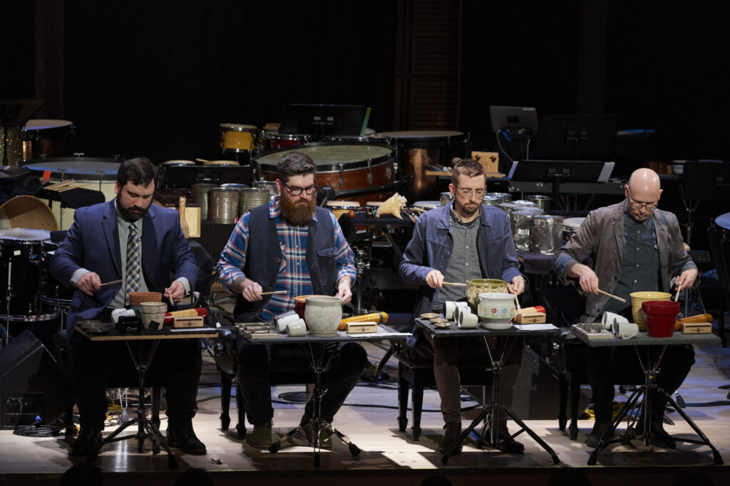 Four musicians performing onstage, each seated at a table with various percussion instruments and objects, including pots and utensils, creating a rhythmic performance.