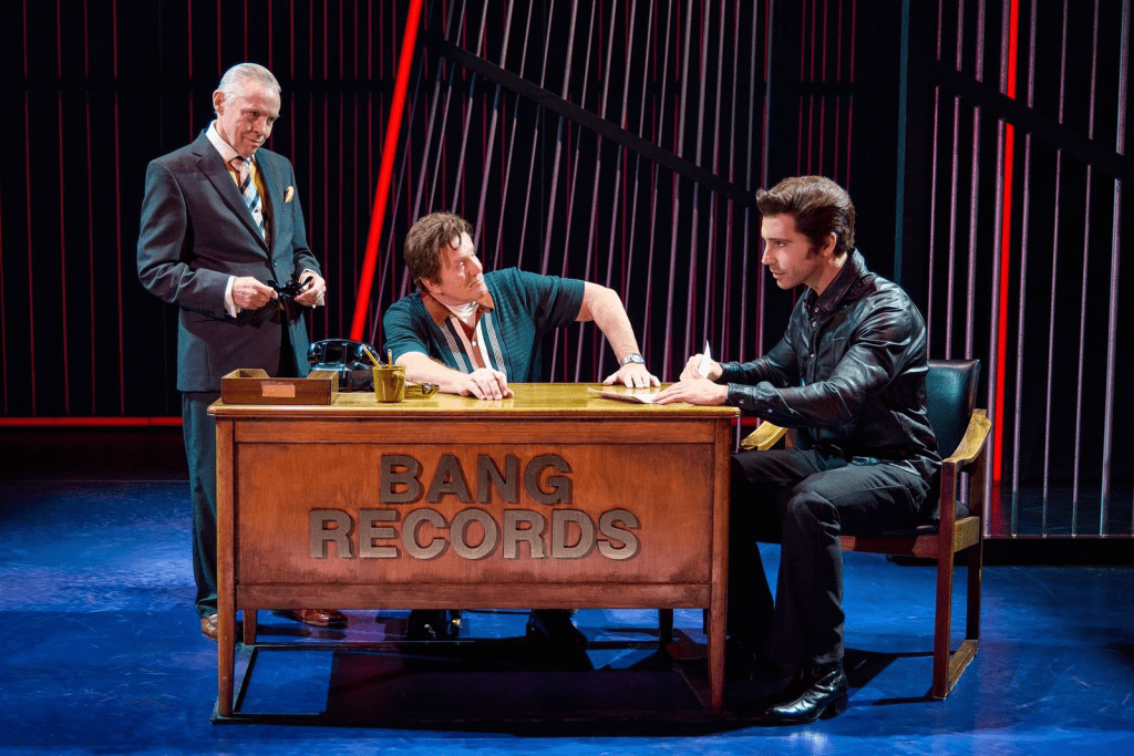 Review: "A Beautiful Noise" Brings Neil Diamond's Music to Life at Salt Lake City’s Eccles Theater 2 A scene depicting three characters in a recording studio setting, with one man in a suit, a woman in the middle, and another man in a leather jacket sitting at a desk labeled 'BANG RECORDS'. The atmosphere is theatrical, highlighting the music industry.