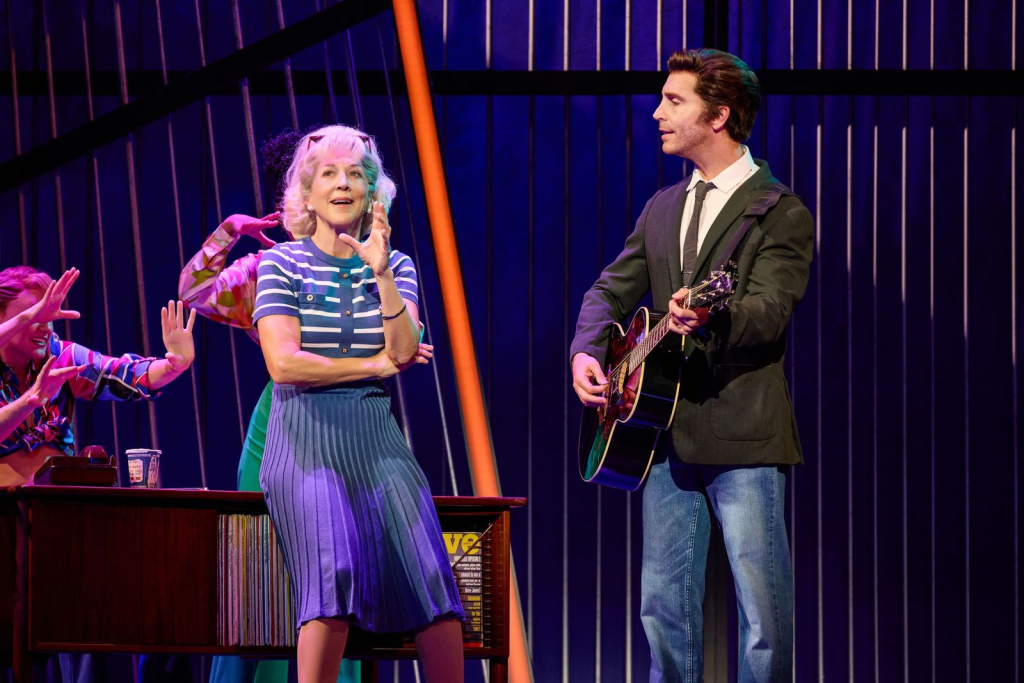 Review: "A Beautiful Noise" Brings Neil Diamond's Music to Life at Salt Lake City’s Eccles Theater 6 A scene from a musical featuring a woman in a striped top and a blue skirt standing beside a desk, while a man in a suit plays guitar. Several background performers are engaging enthusiastically.