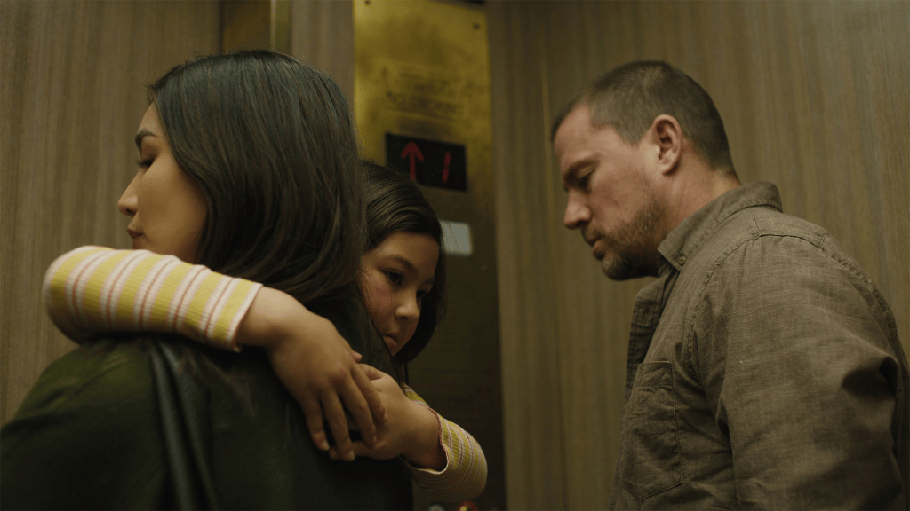 A woman holding a child in an elevator, with a man standing nearby, all appearing serious and contemplative.