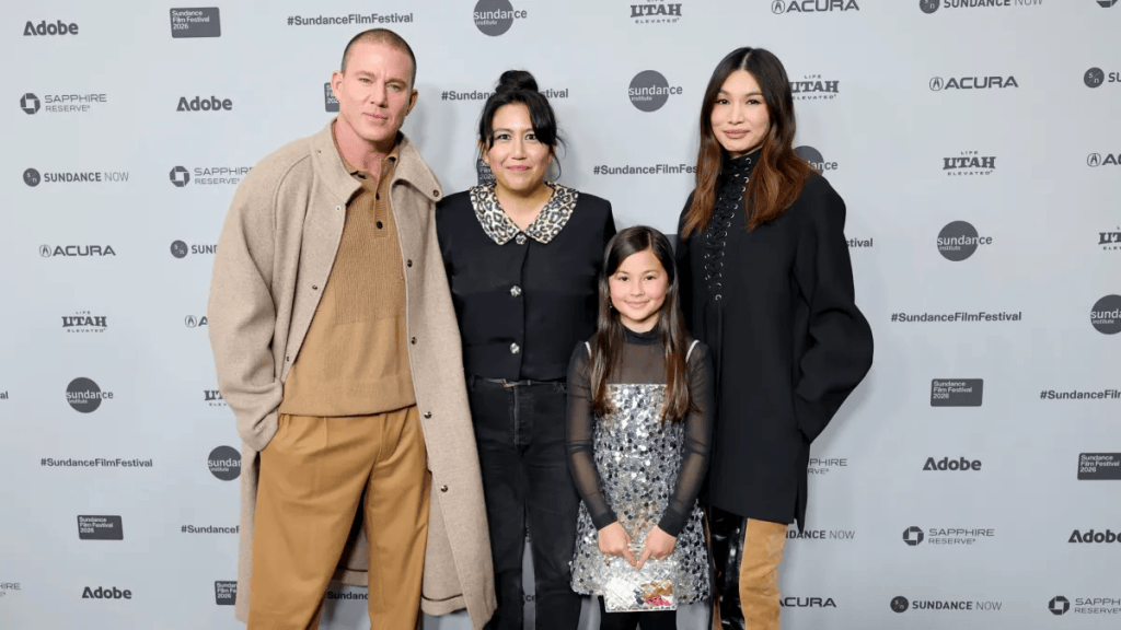 A group of four people, including a man in a brown outfit, a woman in black, a girl in a sparkling dress, and another woman in a stylish black coat, posing together at an event backdrop for the Sundance Film Festival.