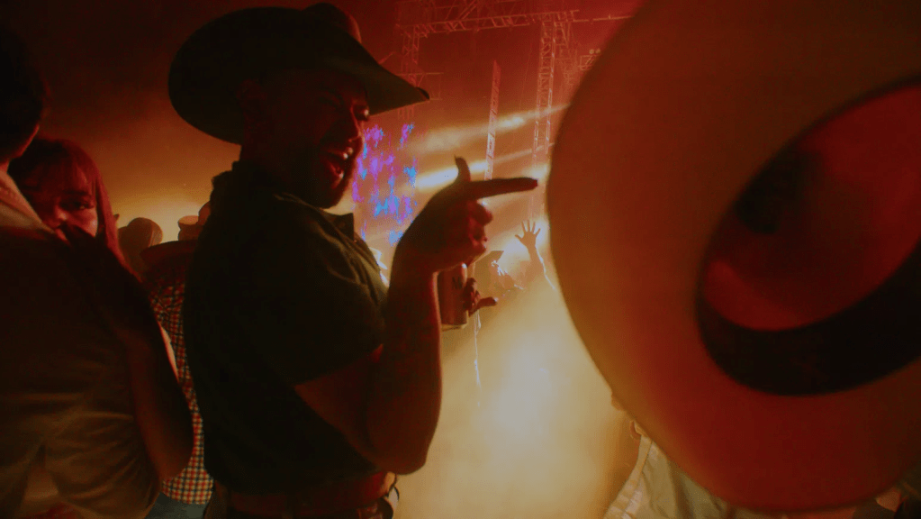 A man wearing a cowboy hat enjoys a lively party atmosphere, pointing and smiling amid a crowd with colorful lights and smoke.