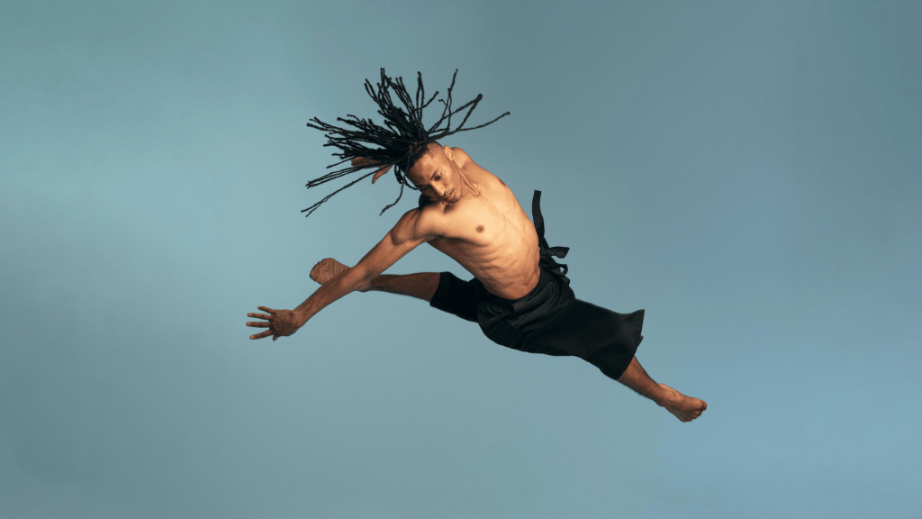 A male dancer in mid-air performing a leap, showcasing athleticism and grace, with long dreadlocks and a bare torso, wearing a black skirt against a light blue background.