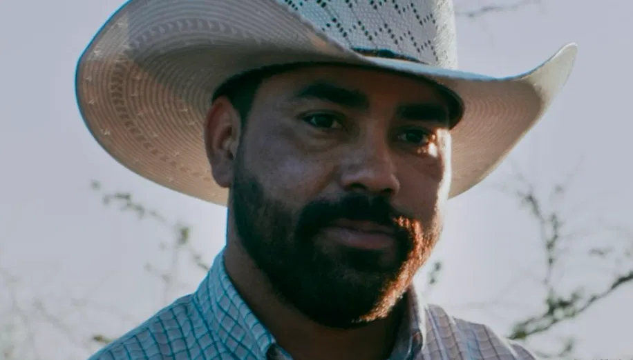 Close-up portrait of a man wearing a cowboy hat and a plaid shirt, standing outdoors with a blurred background of trees.