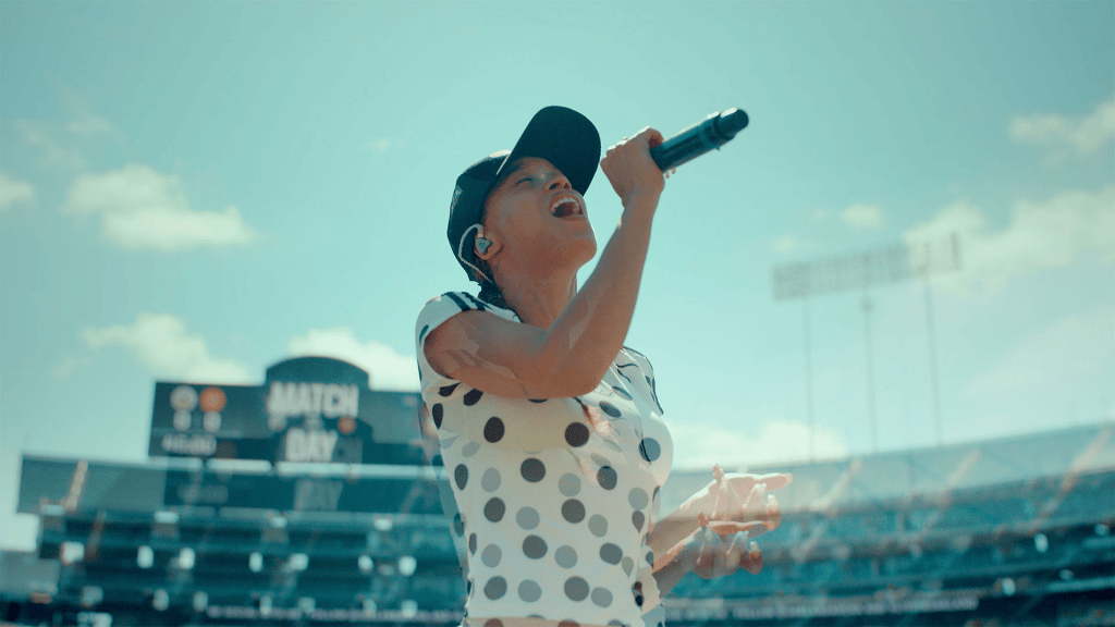 A singer performing on stage at a stadium, holding a microphone and singing passionately, with a blue sky and stadium seats in the background.