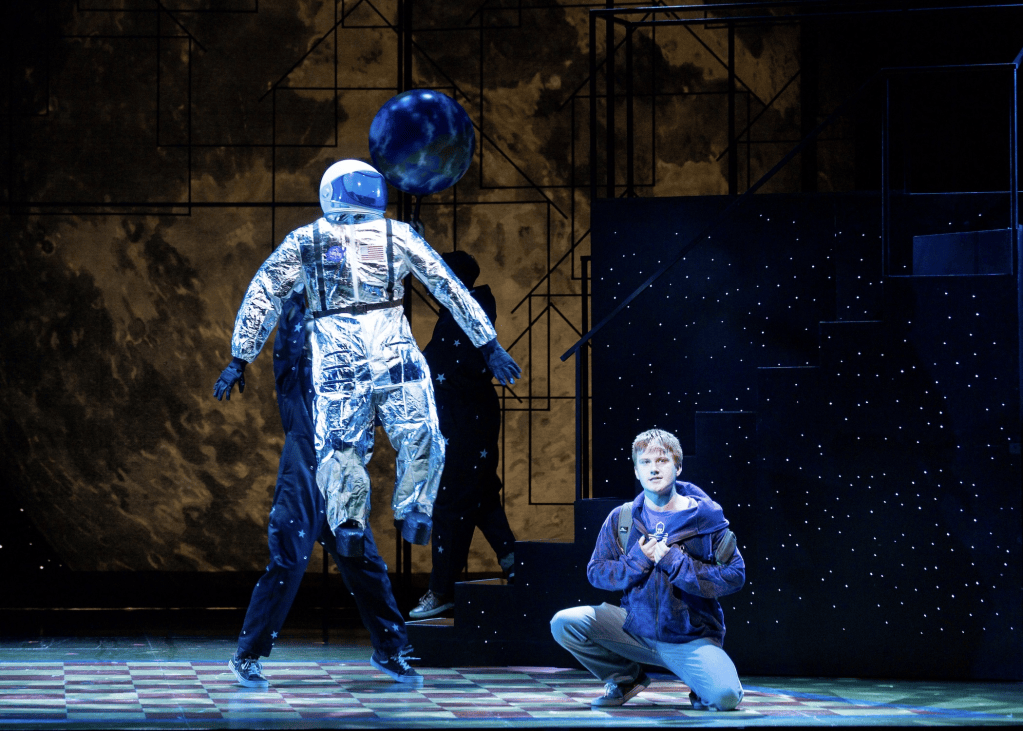 Pioneer Theatre Review: "Ten Brave Seconds" and the Courage of Staying in the Room 2 A theatrical scene featuring a performer in a silver astronaut suit interacting with another character in casual clothing on a stage with a starry backdrop.