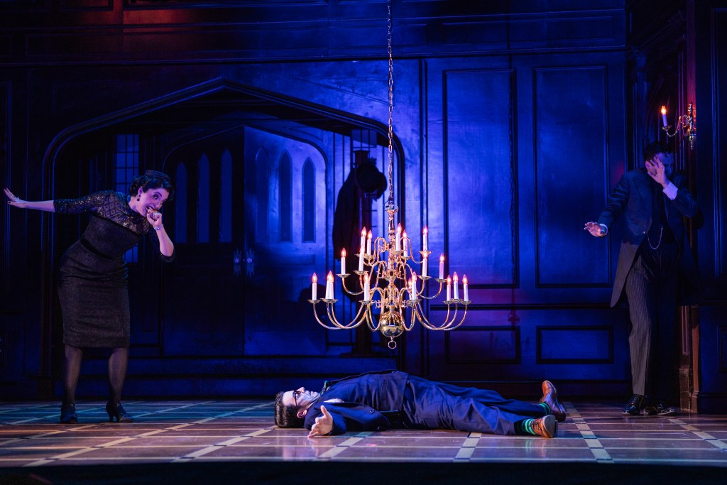 A dramatic stage scene featuring a woman in a black dress reacting with surprise, a man in a suit covering his face, and a man lying on the floor beneath a chandelier.