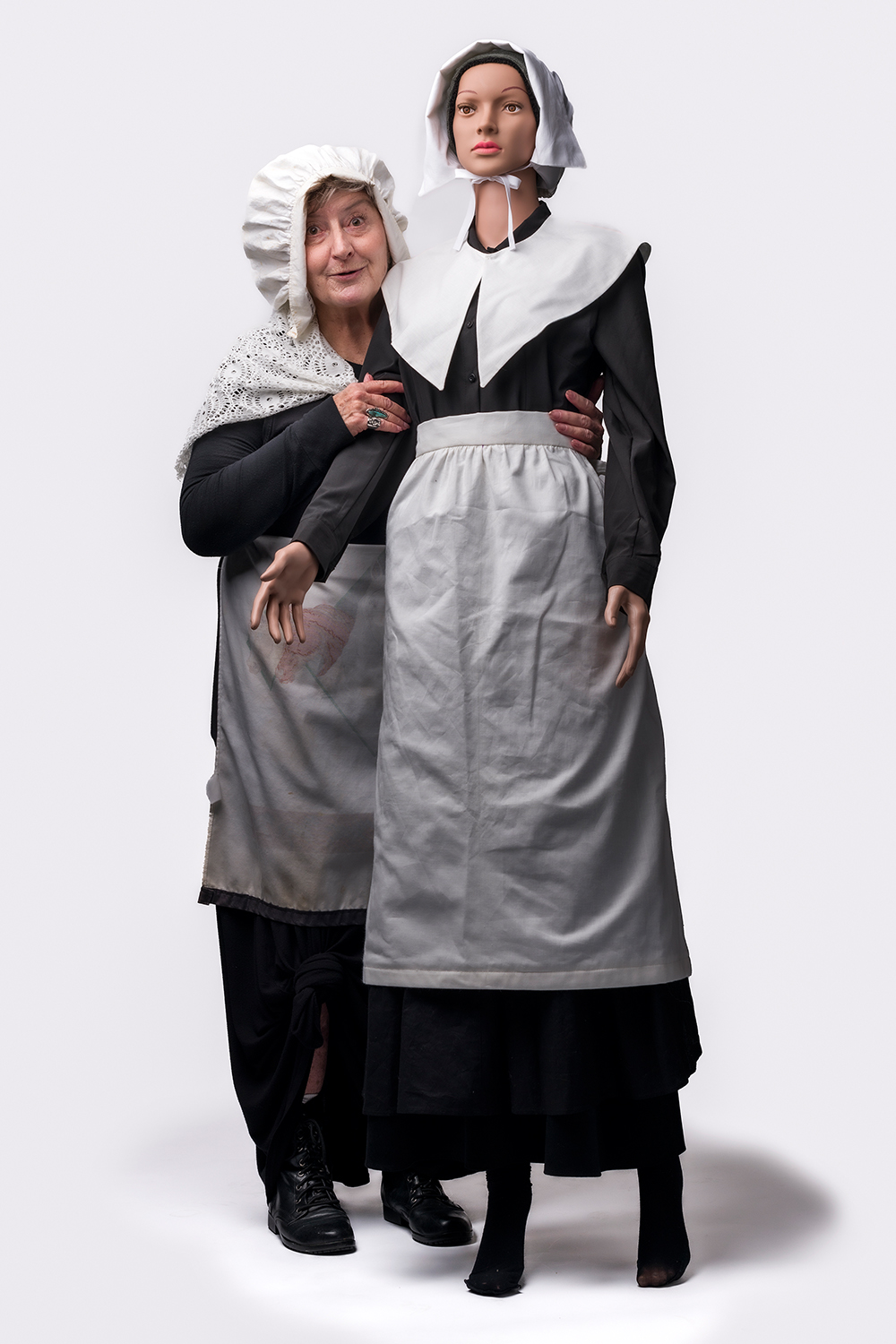 An elderly woman poses with a mannequin dressed in traditional maid attire, both wearing white bonnets, in front of a plain background.