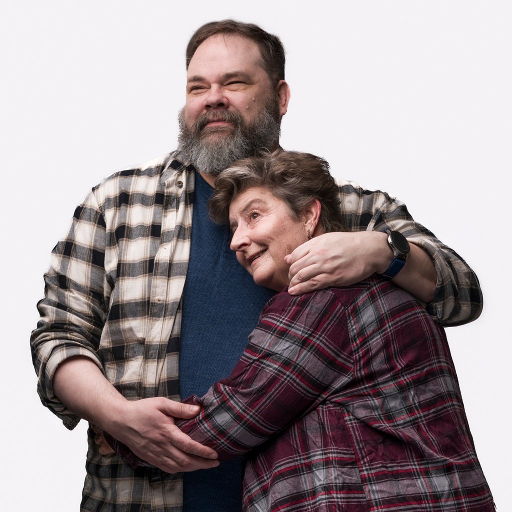 PYG Review: Becky, Nurse of Salem—Returning to the Past to Tell History Differently 7 A man with a beard and a plaid shirt embraces a woman wearing a patterned shirt, both smiling against a plain background.