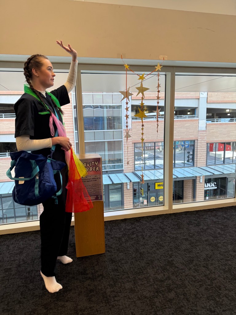 A person in a colorful outfit stands beside a window, playfully interacting with a hanging mobile of stars. They are holding colorful scarves and have a large bag slung over their shoulder.