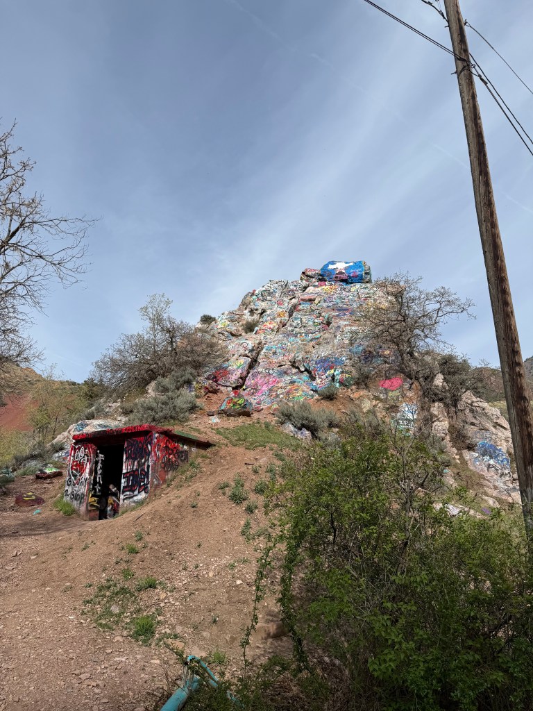 WRTG 4950 Student Group Collaboration on Graffiti Art & Murals in Salt Lake City: Mapping as Rhetoric 5 Colorfully painted rock formation with a small shelter at its base, surrounded by greenery and a clear sky.