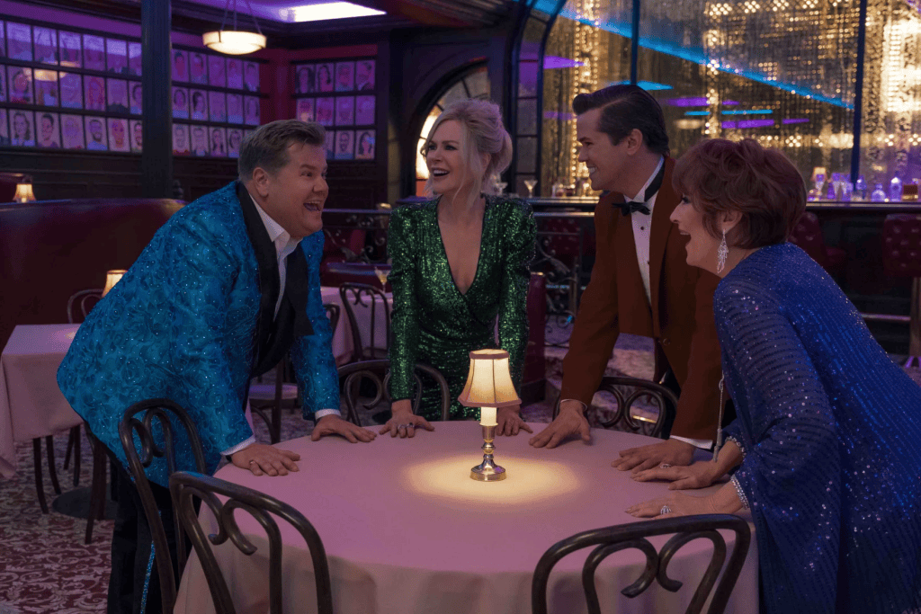 Review: The Prom at Ziegfeld Theater Turns Laughter into a Powerful Reflection on Belonging 2 Four people in colorful, sparkling outfits engage in lively conversation around a table in a dimly lit restaurant, featuring a small lamp.