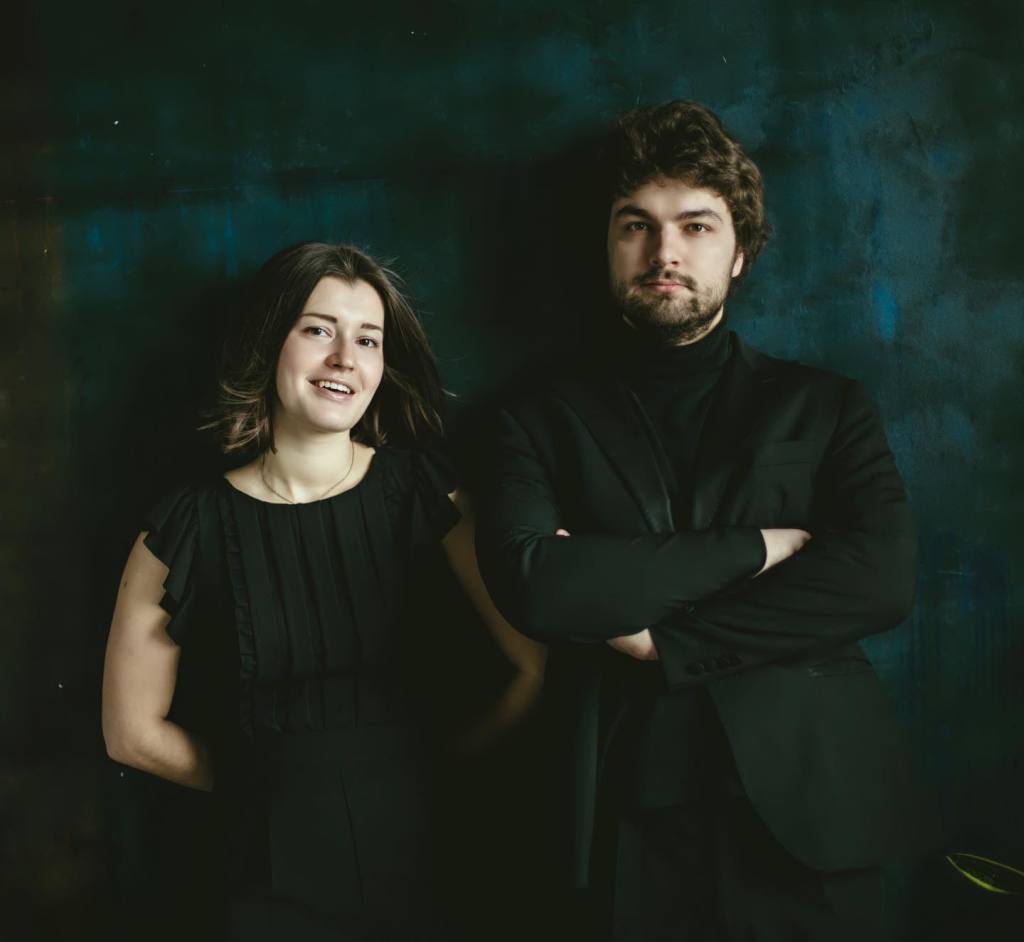 Lukas Geniušas and Anna Geniushene Return to Bachauer: “Heart to Heart” with Rachmaninoff, Copland, and Adams 6 A man and woman standing side by side against a dark textured background. The woman is smiling, wearing a black dress with ruffled sleeves, while the man has a serious expression and is dressed in a black suit.