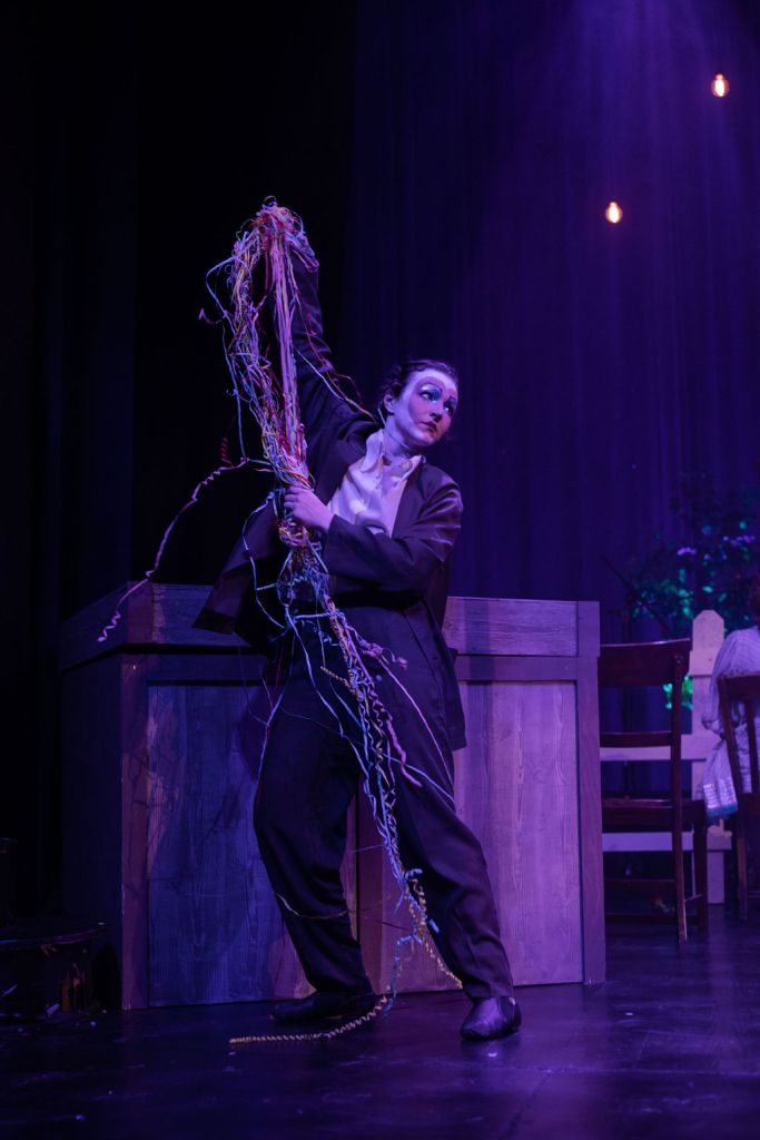 A performer in a dark suit holds a bundle of colorful streamers while standing on stage, with a wooden table and chairs in the background. The stage is illuminated with purple lights.