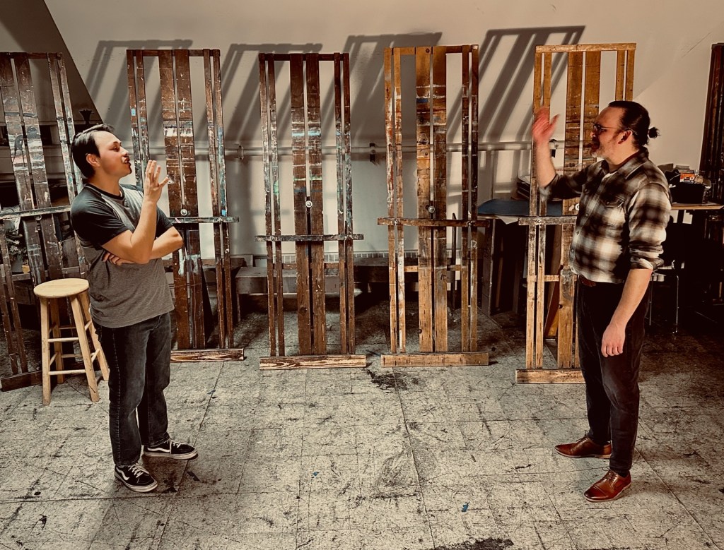 RED Review: Control, Color, and the Cost of Creation 2 Two men engaged in conversation in an art studio, with wooden easels in the background.