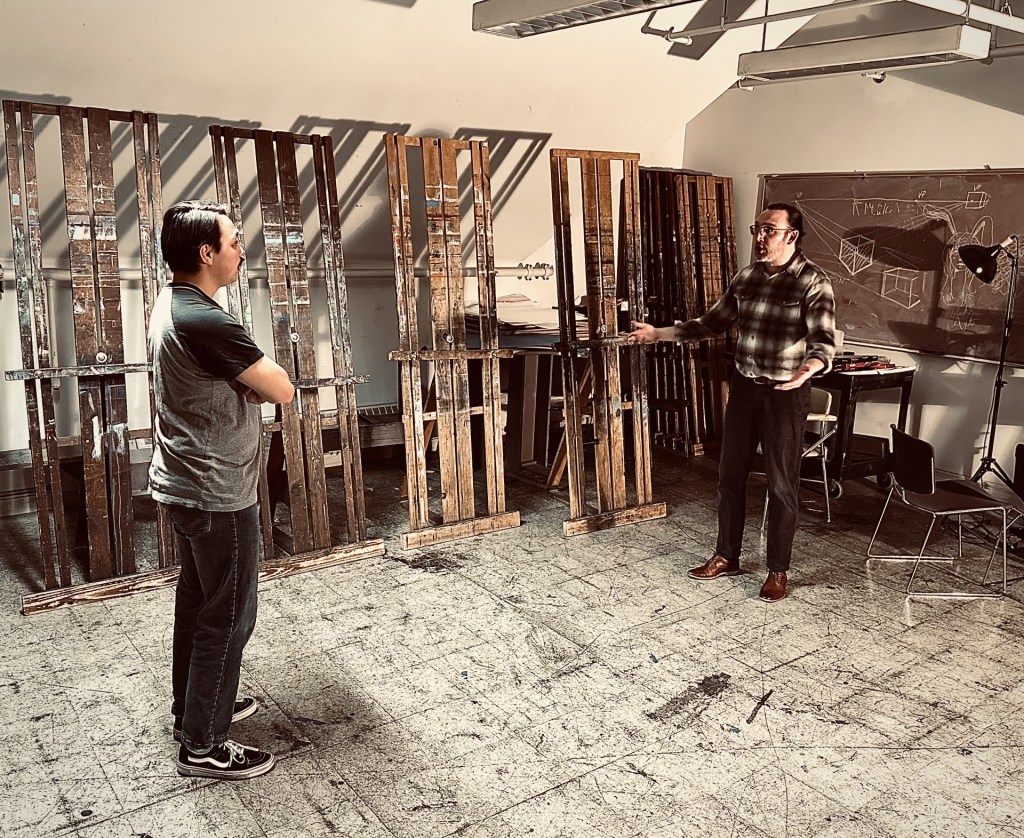 RED Review: Control, Color, and the Cost of Creation 5 Two men engaged in a discussion in an art studio, surrounded by easels and a chalkboard with sketches.