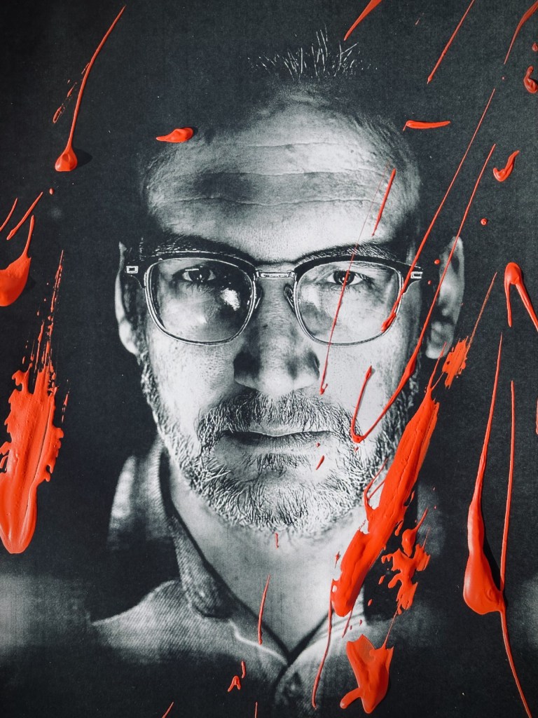 A close-up black and white portrait of a man wearing glasses, with streaks of red paint across the image.