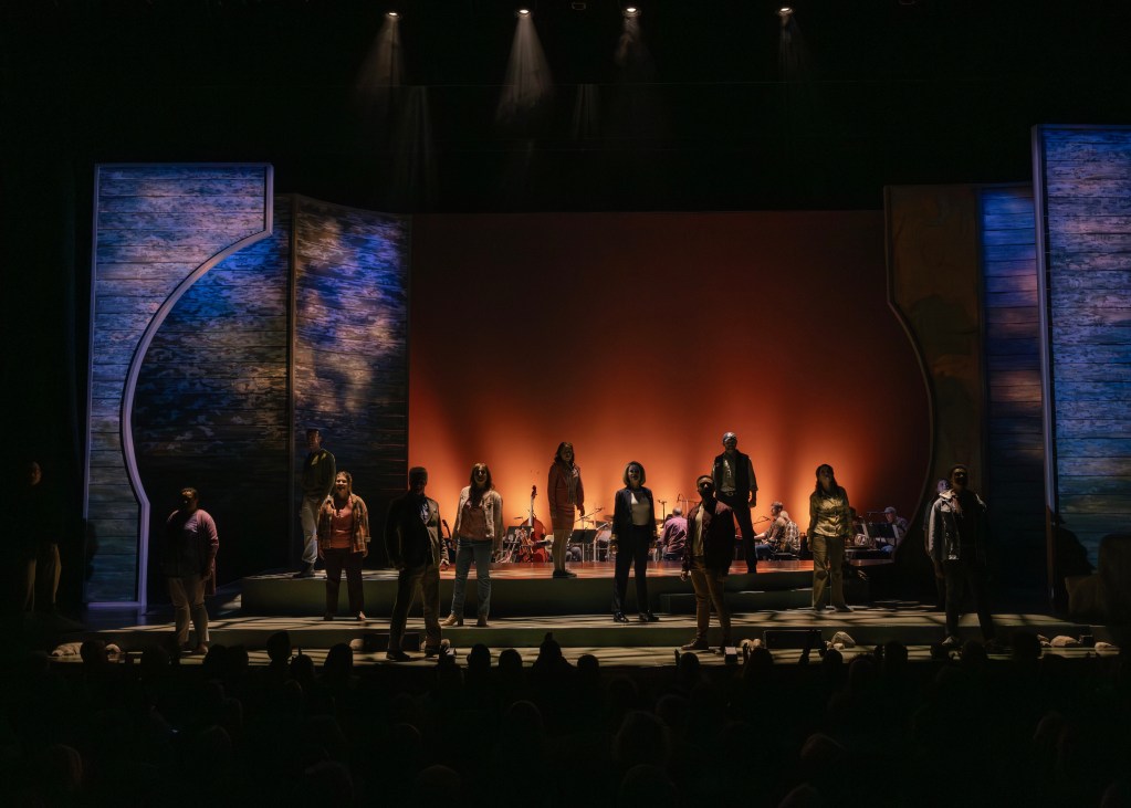 A theater performance scene with actors on stage under dramatic lighting, featuring a vibrant orange backdrop and wooden set pieces.