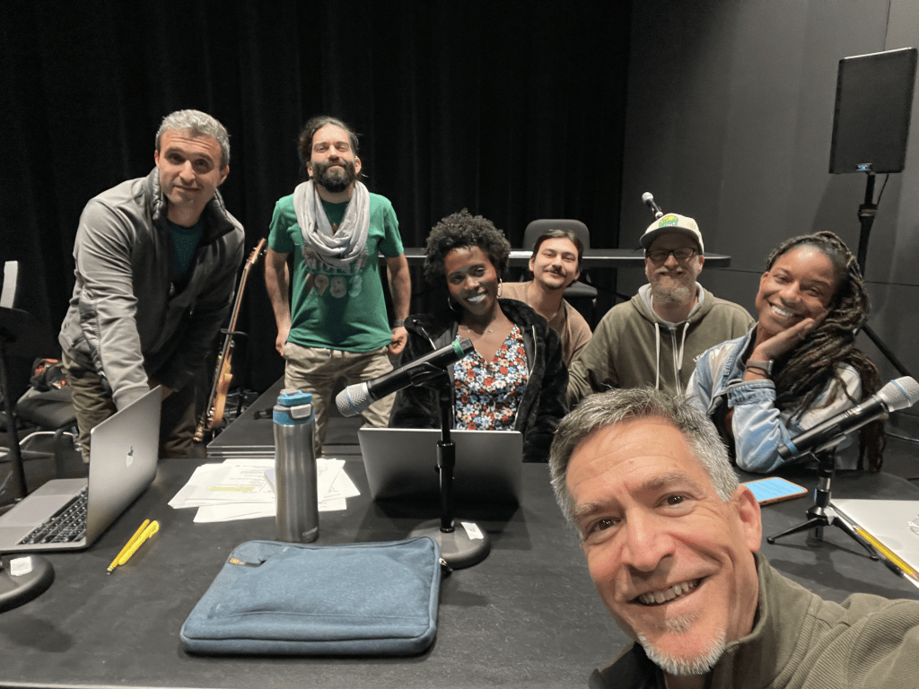 At the Kitchen Table: Dan Froot & Company’s Arms Around America and Theatre as an Ongoing Practice 2 A group of seven people posing together in a recording studio setting, with microphones and laptops on the table.