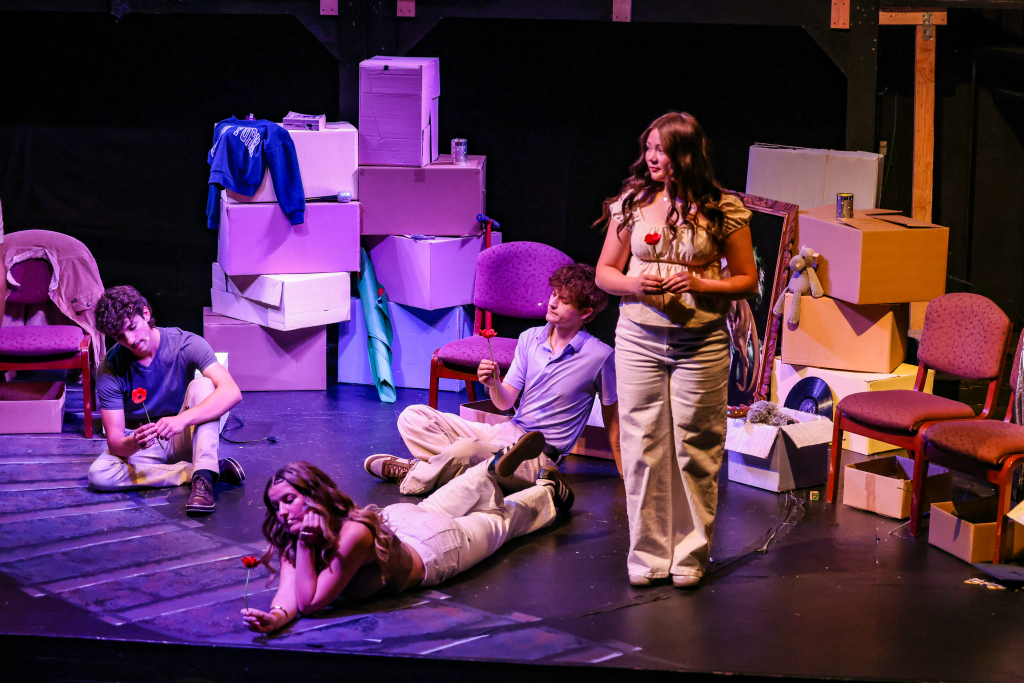 The Keeping Place by Colton Kraus: Memory, Performance, and the Responsibility of Holding 4 A theatrical scene featuring four actors on stage. Two actors are seated on the ground, one holding a flower, while another stands and holds a flower, dressed in casual outfits. The background includes various boxes and chairs, creating a playful set design.