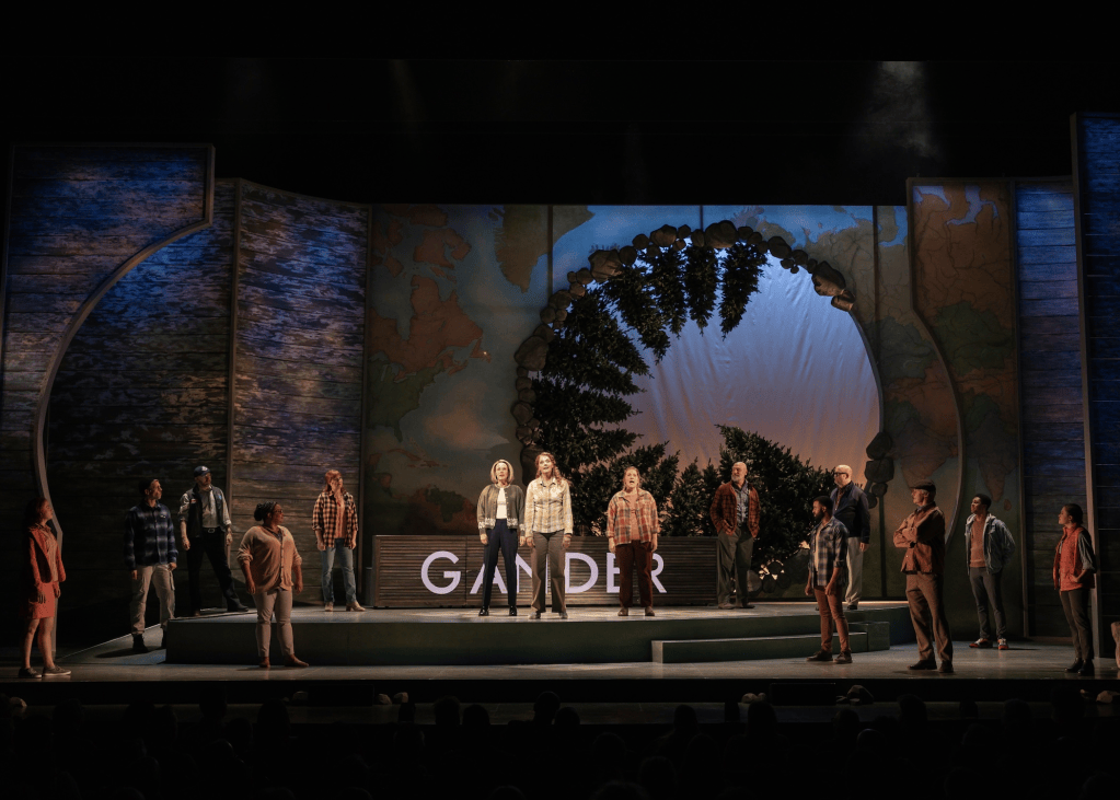 A theatrical performance featuring a diverse cast of actors on stage, with a backdrop depicting a large circular opening surrounded by trees and a map. The word 'GANDER' is prominently displayed on stage.