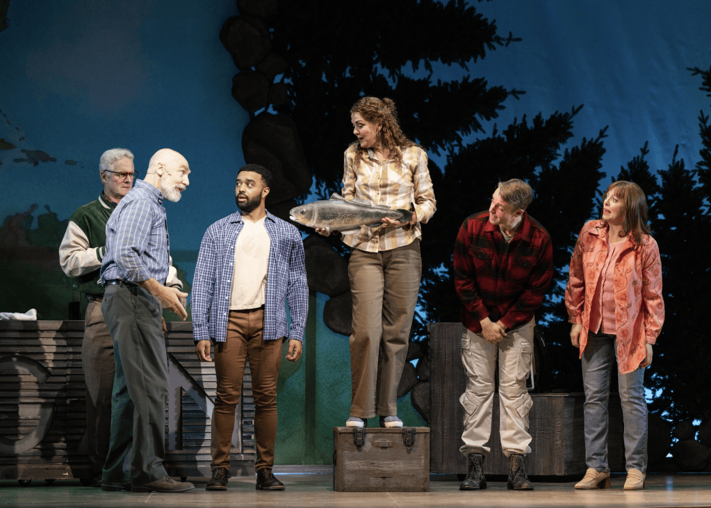 A scene from a theatrical performance featuring five actors in casual outdoor attire, interacting in a rustic setting. One actor holds a large fish, while the others engage in conversation, expressing various emotions.