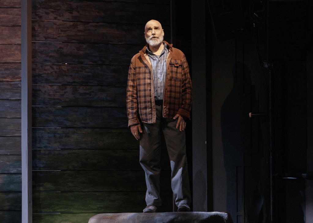 A man with a gray beard and balding head stands on a platform, wearing a plaid shirt and a brown jacket, in front of a wooden backdrop.