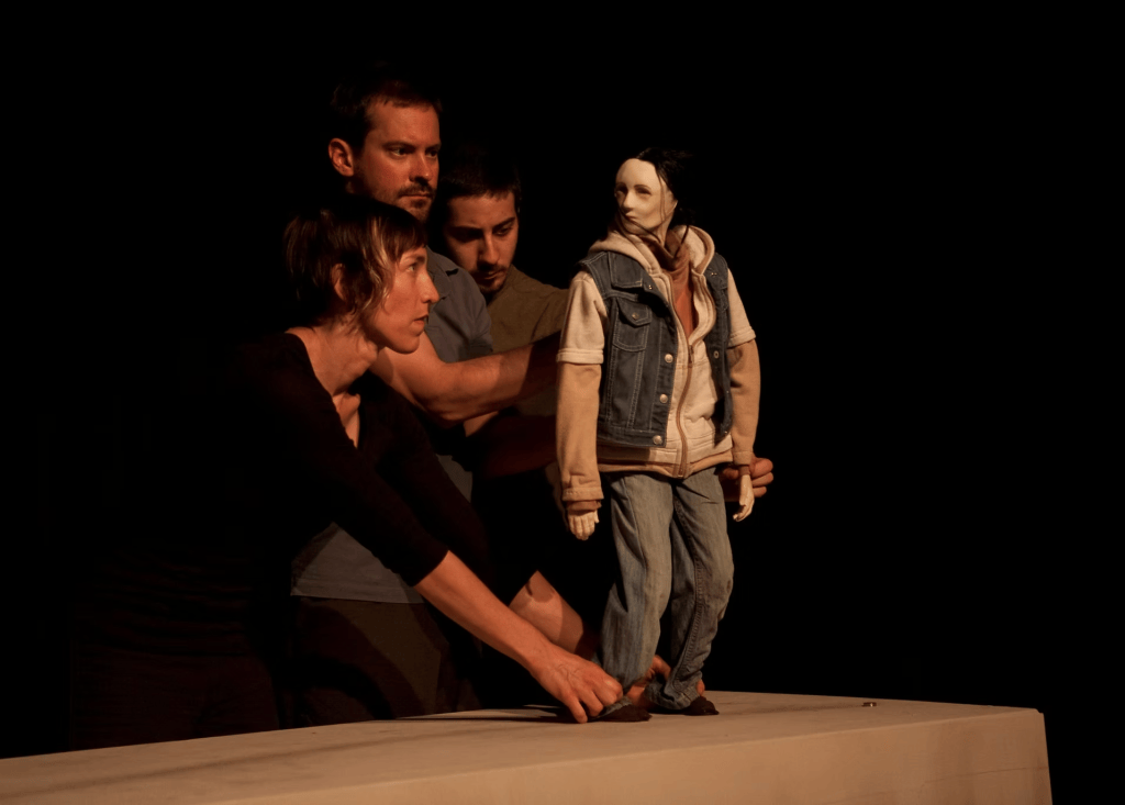 At the Kitchen Table: Dan Froot & Company’s Arms Around America and Theatre as an Ongoing Practice 3 Four performers manipulate a puppet resembling a young person, dressed in a denim jacket and jeans, on a table in a dimly lit space.