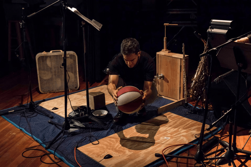 At the Kitchen Table: Dan Froot & Company’s Arms Around America and Theatre as an Ongoing Practice 5 A man kneeling on a wooden floor, holding a basketball in a dimly lit space surrounded by musical instruments and equipment.