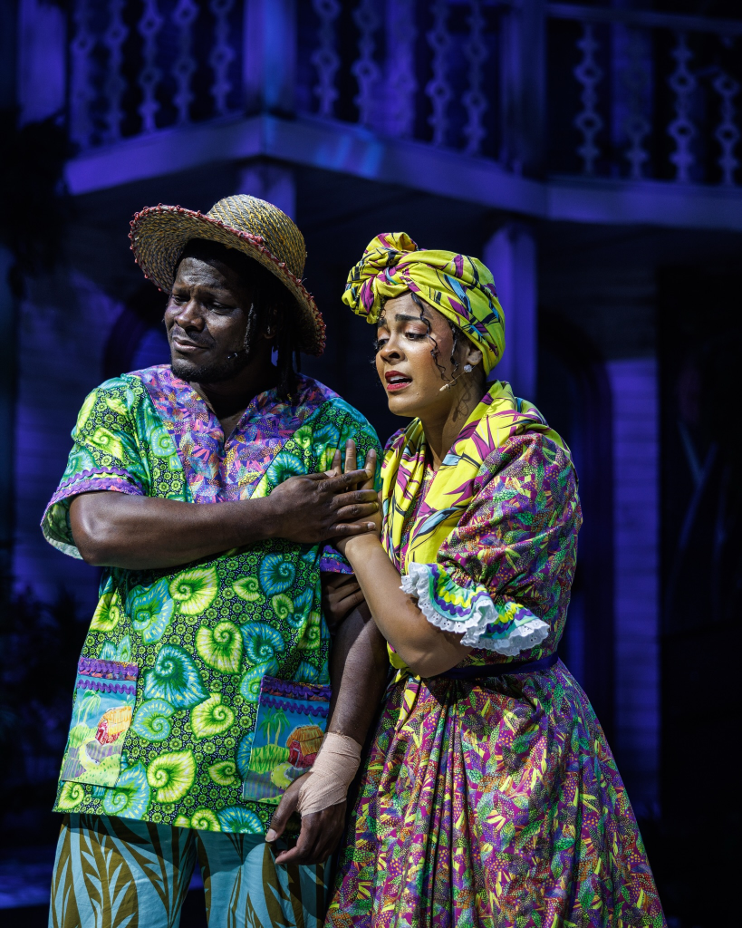 Once on This Island at The Ruth: Storytelling as Survival Through Music and Memory 8 A male and female performer on stage, both dressed in colorful, patterned attire. The man, wearing a straw hat and vibrant shirt with green and blue designs, appears to comfort the woman, who is in a bright, floral dress with a matching head wrap. They are set against a theatrical backdrop.